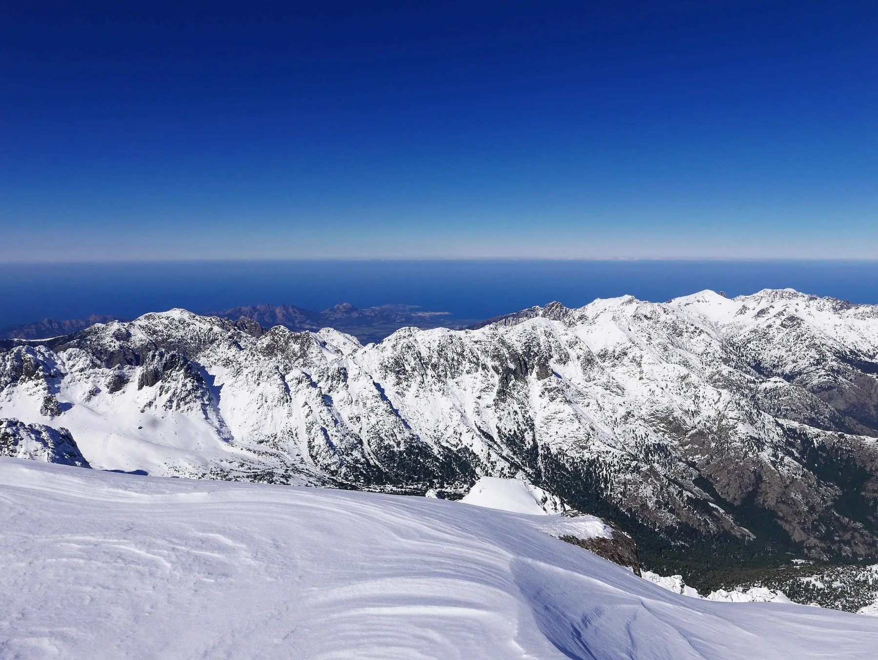 Panorama des montagnes enneigées du Monte Cinto, avec la mer à l’horizon, pendant l’Alpine Line en Corse.